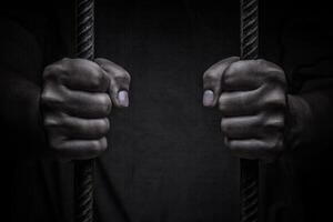 de cerca en manos de hombre sentado en celda. hombre detrás cárcel barras en negro antecedentes foto