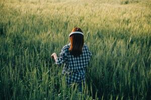 joven bonito mujer en rojo verano vestir y Paja sombrero caminando en amarillo granja campo con maduro dorado trigo disfrutando calentar noche. foto