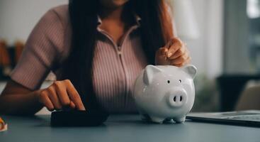 joven asiático mujer ahorro para Jubilación ahorro dinero mediante un cerdito banco y tomando notas en computadora portátil, ahorros concepto. foto