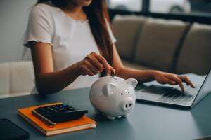 joven asiático mujer ahorro para Jubilación ahorro dinero mediante un cerdito banco y tomando notas en computadora portátil, ahorros concepto. foto