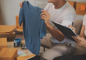 Two Asian blogger showing clothes in front of the camera to recording vlog video live streaming at her shop. Online Shopping Cart notion. on sofa at home photo