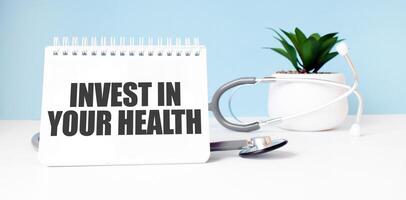 The text INVEST IN YOUR HEALTH is written on notepad near a stethoscope on a blue background. Medical concept photo