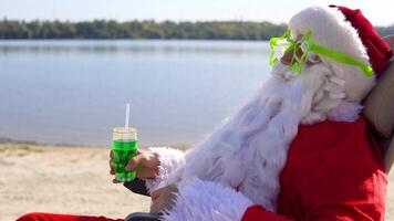 Santa Claus in funny glasses drinks a green cocktail while lying on a sun lounger on the lake beach. Travel and vacation concept video