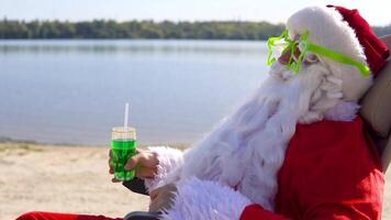 santa claus dentro engraçado óculos bebidas uma verde coquetel enquanto deitado em uma Sol espreguiçadeira em a lago de praia. viagem e período de férias conceito video