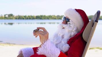 Père Noël dans des lunettes de soleil fume une cigare tandis que mensonge sur une Soleil fainéant sur une Lac plage video