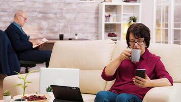 Zoom in shot of elderly age woman with glasses taking a sip of coffee browsing on phone. Elderly age man reading a book in the background. video