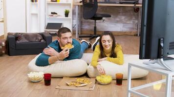 Bearded man drinking soda and sitting on pillows for the floor with his attractive girlfriend watching tv while the cat is sleeping in the background. video