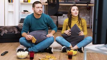 Disappointed couple sitting on the floor while watching sport on tv and eating pizza with their cat sleeping. video