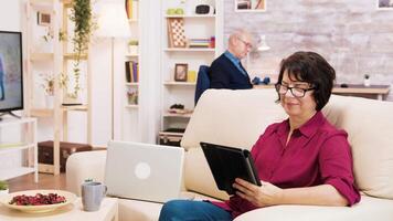 Zoom in shot of elderly age woman using tablet sitting on coich. Senior man reading a book in the background. video