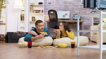 Zoom in shot of young couple sitting on pillows for the floor eating pizza and watching tv while their cat in sleeping in the background. video