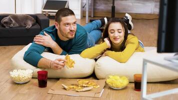 atractivo Pareja relajante en almohadas para el piso acecho televisión y comiendo Pizza con su gato dormido en el antecedentes. video