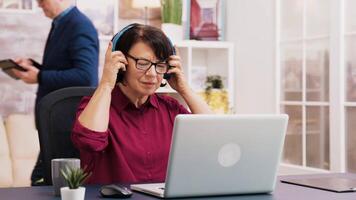 Old woman enjoying a cup of coffee while working on laptop with headphones on her head. Elderly man using tablet in the background. video