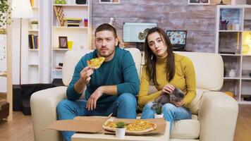 Beautiful young woman sitting on couch with her boyfriend watching tv, eating pizza and playing with their cat. video