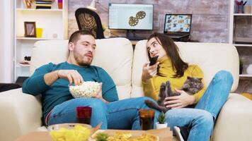 Bored girl sitting on the couch with the cat in her lap and boyfriend next to her is using tv remote control. video