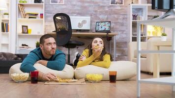 Happy caucasian couple eating pizza sitting on pillows for the floor watching tv while their cat is relaxing in the background. video