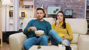 jong paar ontspannende Aan sofa aan het kijken TV terwijl aan het eten popcorn en chips. video