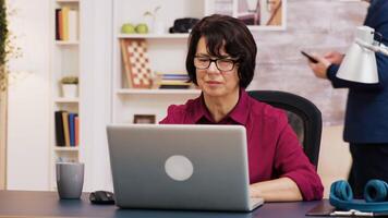 pensionato donna Lavorando su il computer portatile nel vivente camera mentre sua marito è utilizzando tavoletta nel il sfondo. video