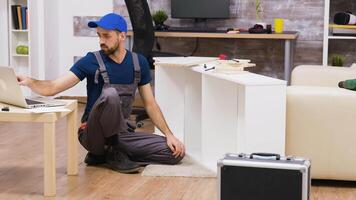 Professional worker in overalls consults furniture assembly instructions from laptop. Worker wearing a cap. video