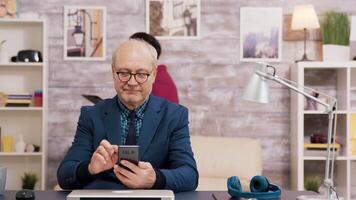 Elderly man with glasses browsing on mobile phone in living room. Woman walking and using tablet in the background. video