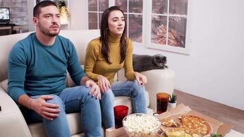 chateado jovem casal enquanto assistindo uma jogos em televisão. gato sentado em a sofá. pizza, refrigerante, e Pipoca em café mesa. video