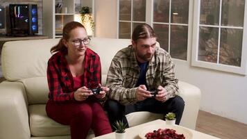 Man and woman sitting on sofa playing video games using wireless joysticks.