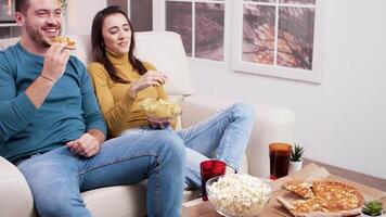 caucásico joven Pareja sentado en sofá comiendo Pizza mientras acecho televisor. palomitas de maiz y soda en café mesa. video