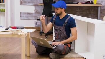 Furniture assembly worker wearing overalls searching instructions using his laptop. Worker holding screwdriver. video