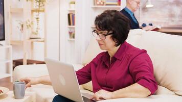 Side view of retired woman typing on laptop sitting on sofa. Husband in the background reading a book. video