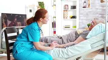 Female assistant from nursing home taking the hand of old lady lying in hospital bed and talking with her video