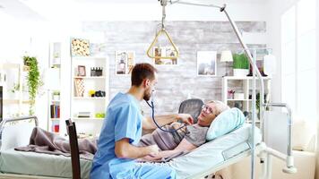masculino enfermero comprobación el latidos del corazón de un enfermo antiguo dama acostado en hospital cama en brillante y acogedor enfermería hogar video