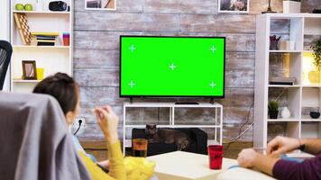 Back view of young couple sitting on chairs looking at tv with green screen, eating popcorn with the cat watching them. video