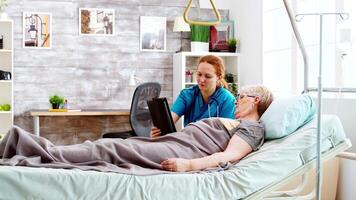 kvinna vårdgivare hjälper ett gammal Inaktiverad kvinna liggande i sjukhus säng till använda sig av en digital läsplatta st. ljus rum med stor fönster video