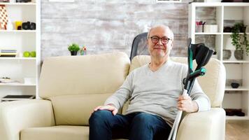 Portrait of elderly retired man smiling to the camera in cozy nursing home, he has crutches next to the sofa video