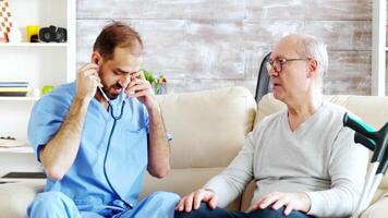 Revealing shot of young male nurse listening to old retired man heartbeat in a bright and cozy nursing home. Caregiver and social worker video