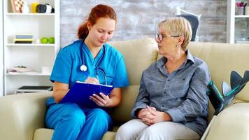 femmina medico con rosso capelli assunzione Appunti su appunti mentre parlando con anziano donna su il divano nel assistenza infermieristica casa video