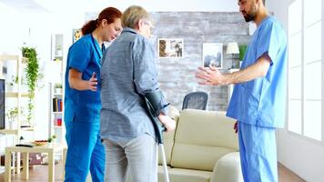 Male doctor and his assistant in nursing home helping senior woman who walk in in crutches to sit on couch video