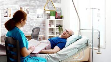 Old retired sick man lying in hospital bed in a nursing home while a nurse is reading him a book video