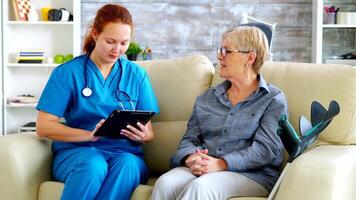 Female doctor holding tablet computer in nursing home talking with senior woman on the couch video