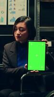 Vertical Video Law agent in wheelchair shows greenscreen display on tablet, using isolated mockup layout on device in disability friendly archive room. Investigator holding gadget with chromakey screen. Camera A.