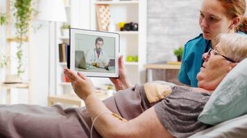 Old disabled woman lying in hospital bed having an online video call with a doctor. A nurse is next to her