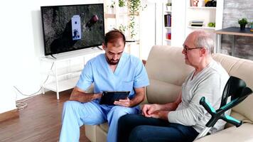 Experienced doctor taking notes on tablet computer while talking with old man in retirement home. video