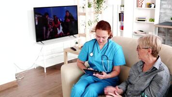 hembra enfermero participación tableta ordenador personal en Jubilación hogar hablando con mayor mujer en el sofá video