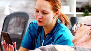 Close up shot of nurse helping an elderly disabled woman to use a smartphone. Bright room with big windows video