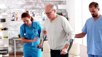 jovem masculino médico e mulher ajudando velho homem dentro enfermagem casa para andar com muletas. video