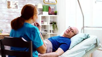 antiguo enfermo hombre en un hospital cama obtiene su latido del corazón comprobado por un joven caucásico enfermero. brillante habitación con grande ventanas video