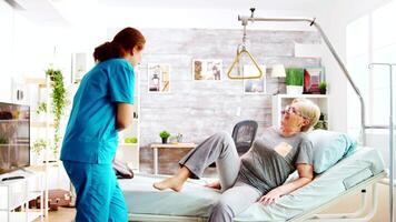 Female nurse helping an old lady to go to bed and covers her with a blanket in a nursing retirement home with big windows video