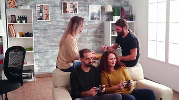 Bearded man with long hair eating chips in living room while his friends are playing video games with wireless controller.