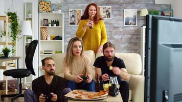 Young man eating chips while playing video games on tv with his friends sitting on the couch. Girl holding a beer bottle.