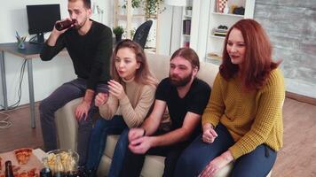 grupp av fyra vänner tittar på en film i levande rum Sammanträde på de soffa dricka öl och äter pizza. video