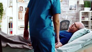 Nurse taking a seat near an old sick man who lies in hospital bed in a nursing retirement home video